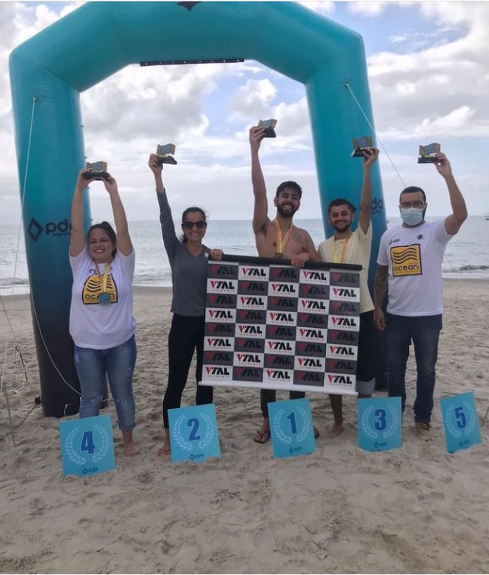 #Pracegover foto: na imagem há cinco pessoas, placas de números, o mar e areia