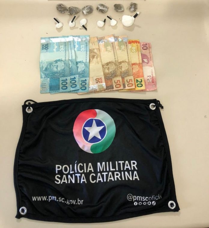 #PraCegoVer Na foto, uma bandeirola com o símbolo da Polícia Militar, drogas e dinheiro apreendidos