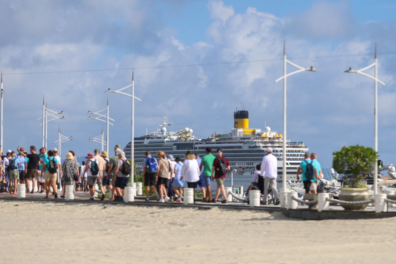 Foto mostra fila de passageiros ansiosos para ocupar seus lugares no cruzeiro marítimo. Navio ancorado ao fundo.