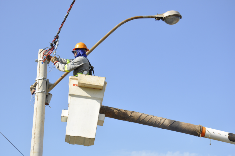 Imagem mostra manutenção em poste de energia com operário içado por equipamento especial vistoriando poste e luminária pública.